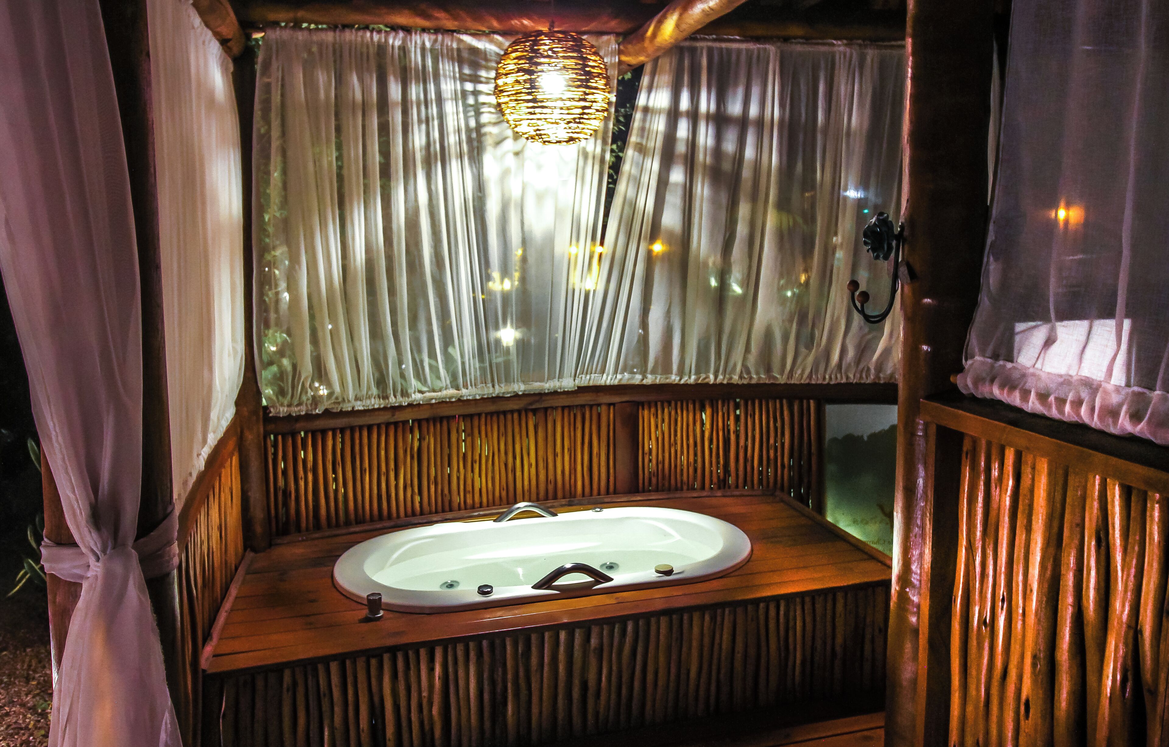 A wooden cabin with a wooden bathtub, a hanging light fixture, and sheer curtains.