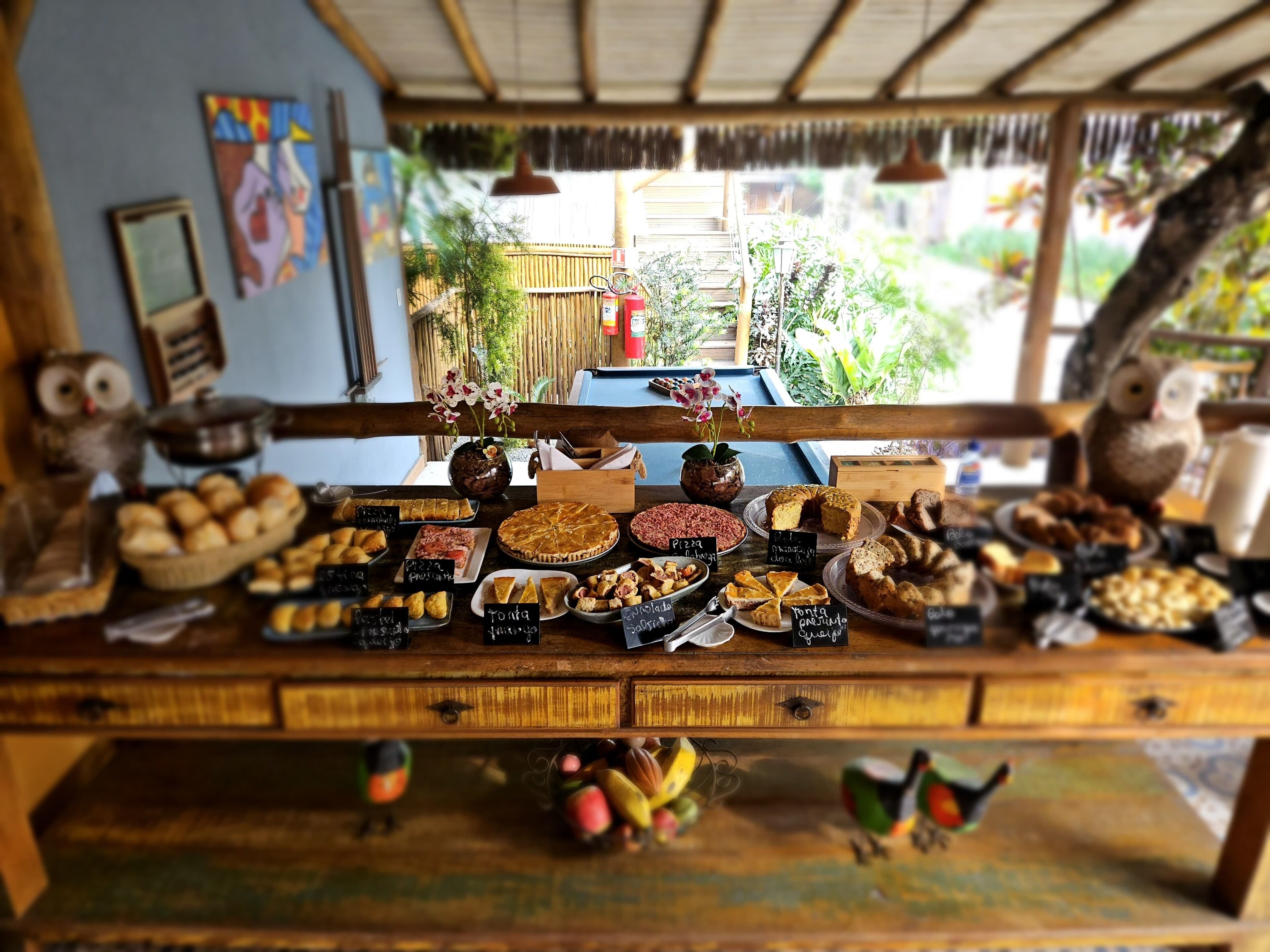 Een houten toonbank met verschillende gebakjes en patisserie, een rustiek interieur en uitzicht op groen.