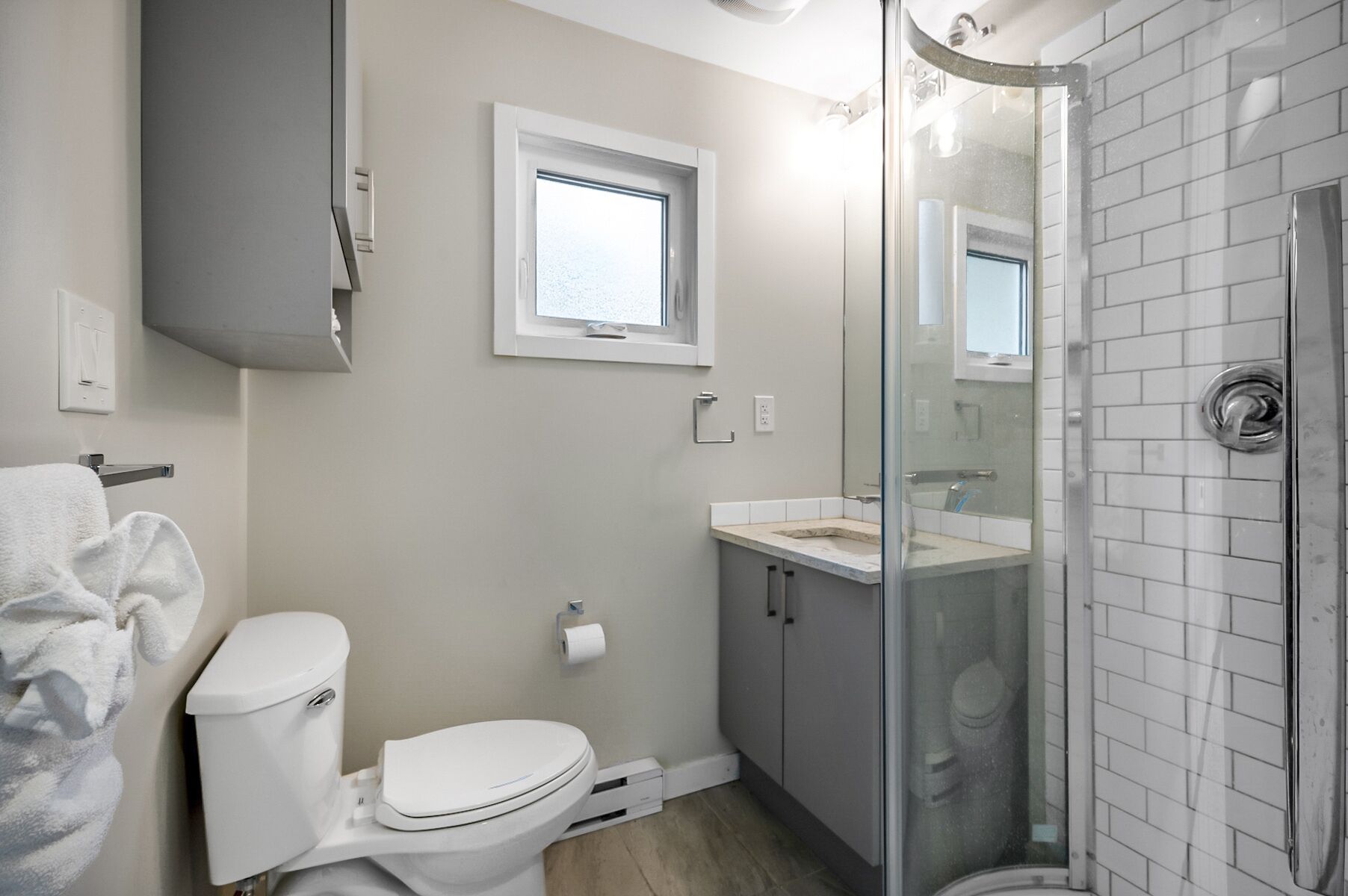 A modern bathroom with a toilet, a glass shower enclosure, and a sink with a window above.