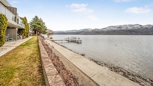 A lakeside property with a dock extending into the water, a grassy area with a stone border, and a building with a balcony overlooking the lake. - Family Friendly Cottage w Amazing View (West Kelowna)