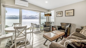 A spacious living room with a large sliding glass door leading to a balcony with a lake view. There are two leather armchairs, a white dining table with chairs, and a patterned ottoman. - Family Friendly Cottage w Amazing View (West Kelowna)
