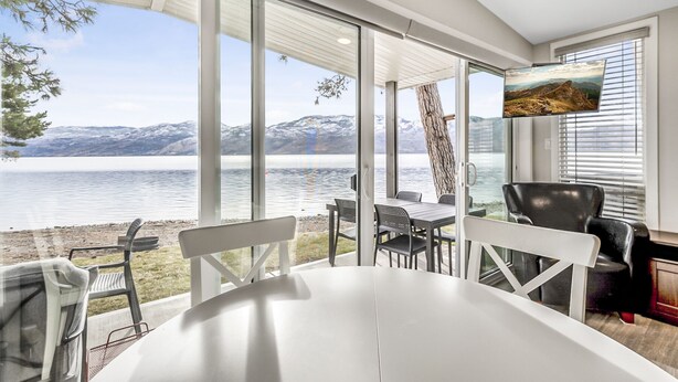 A modern dining area with a large table and chairs, overlooking a lake with mountains in the background. - Family Friendly Cottage w Amazing View (West Kelowna)