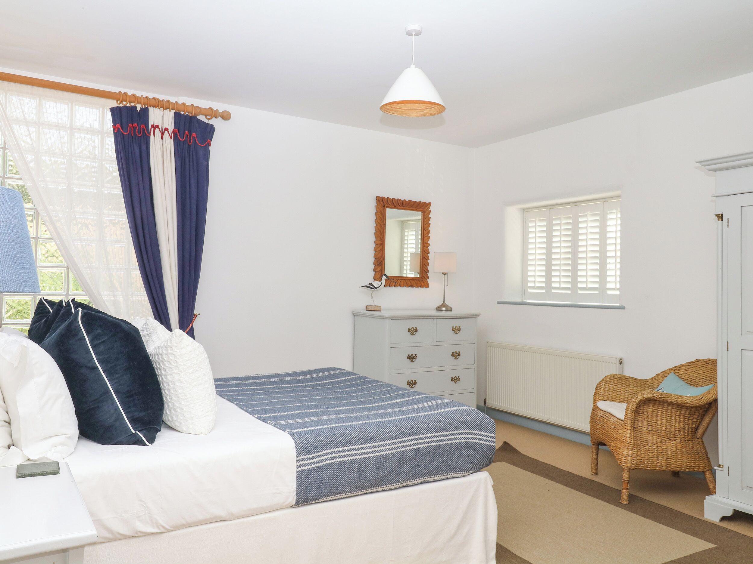 A bedroom with a bed, a dresser, a chair, a window with shutters, and a hanging light fixture.