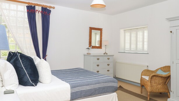 A bedroom with a bed, a dresser, a chair, a window with shutters, and a hanging light fixture. - Burrows (Ottery St Mary)