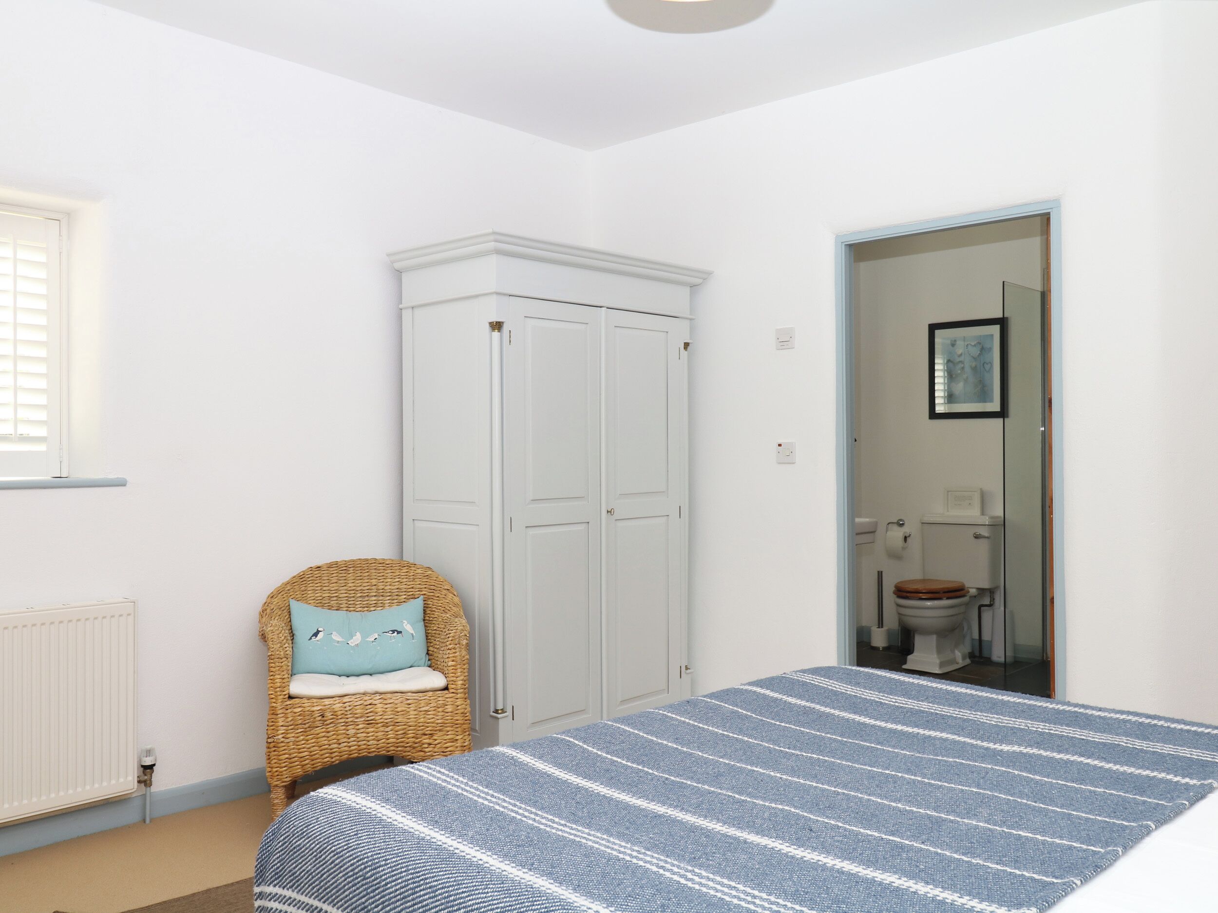 Quarto com cama, guarda-roupa, cadeira de vime e banheiro visível por uma porta aberta.