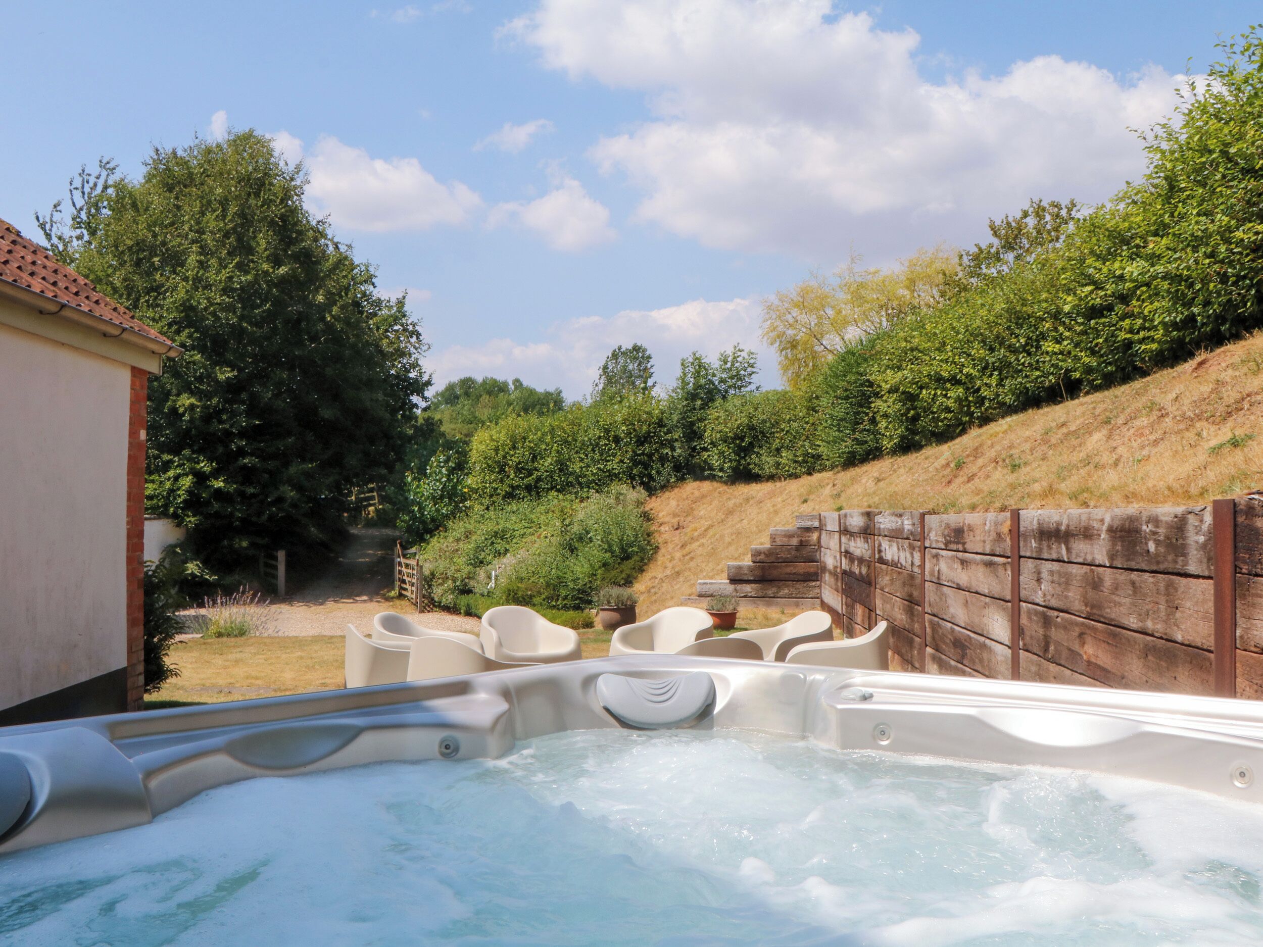 Un espace avec un jacuzzi, entouré d’une clôture en bois, comprenant des sièges extérieurs et un bâtiment visible au loin.