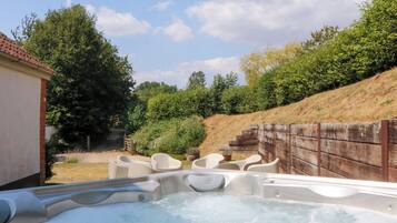 Un espace avec un jacuzzi, entouré d’une clôture en bois, comprenant des sièges extérieurs et un bâtiment visible au loin.