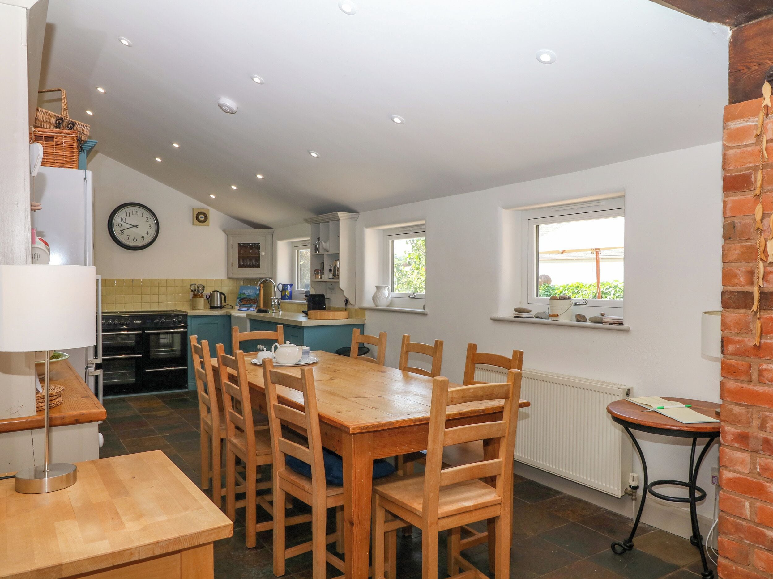 A kitchen with a dining area, wooden chairs, a clock, and a window with a view.