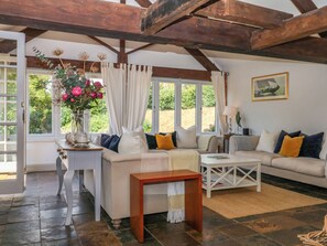 A cozy living room with a sofa, a coffee table, and a wooden bench. There is a window with curtains and a vase of flowers on a side table. - Apple Mill (Ottery St Mary)