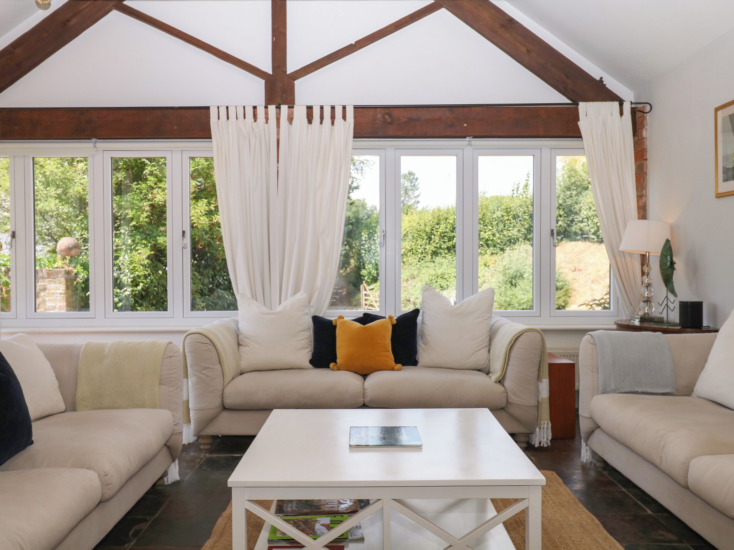 A room with a high ceiling, exposed wooden beams, a beige sofa set, a white coffee table, and large windows with curtains.