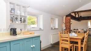 A kitchen with a blue cabinet, a dining table with chairs, and a window with a view of greenery. - Apple Mill (Ottery St Mary)