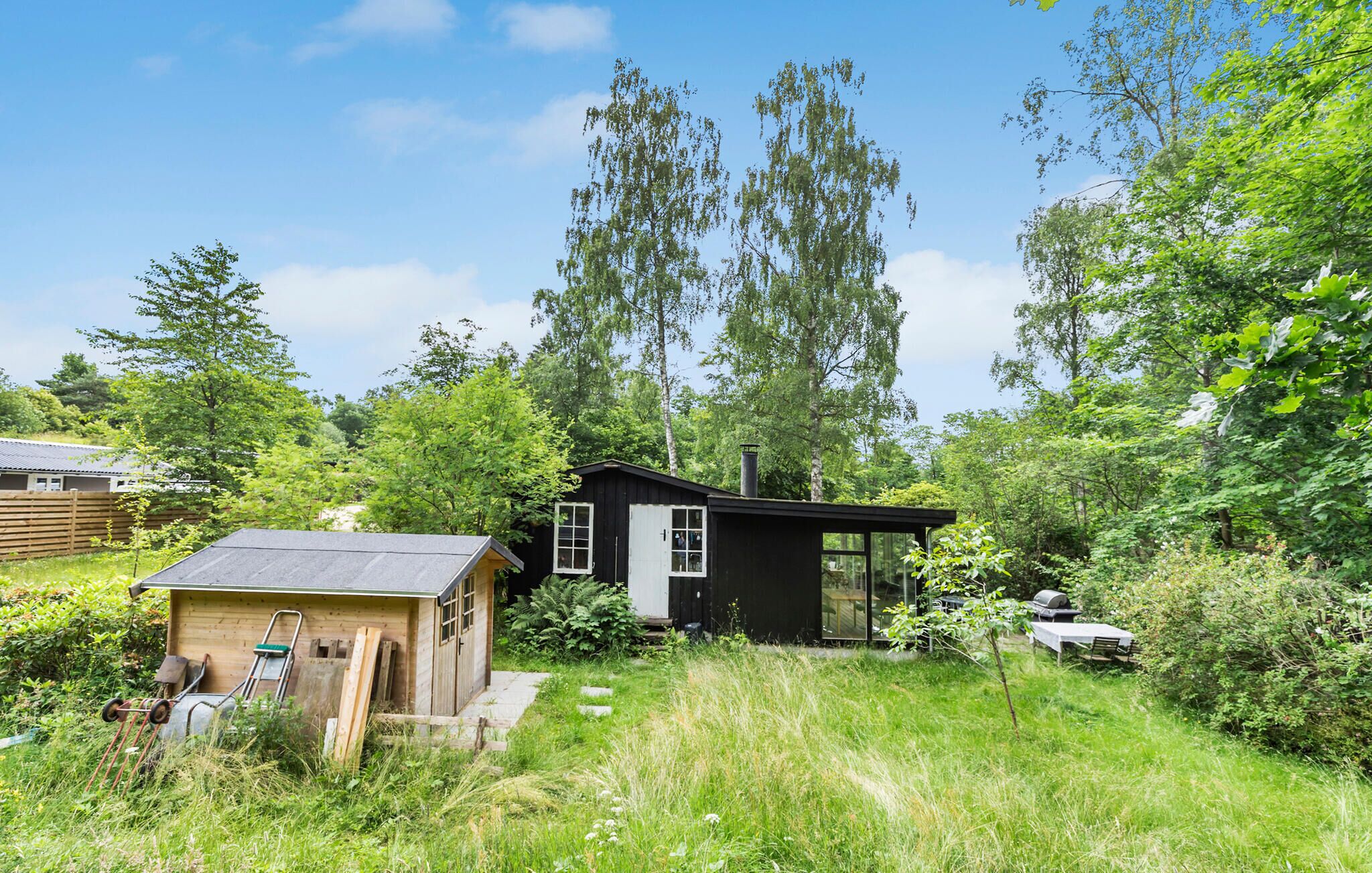 A small, single-story cabin with a sloped roof, surrounded by dense greenery and tall trees.