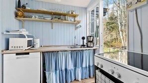 Una cocina con microondas, cafetera y estufa integrados. Hay repisas de madera sobre la encimera con platos y vasos. Una ventana ofrece vista a los árboles.