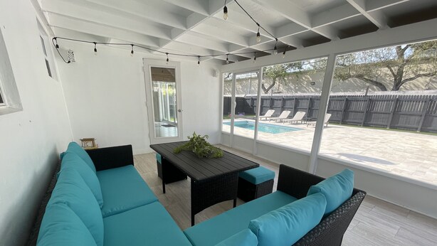 Un patio couvert avec une table et des chaises noires, un canapé turquoise doté de coussins, et une vue sur une piscine.