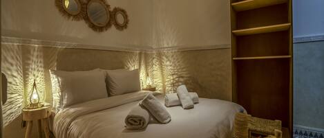 A neatly made bed with white linens and rolled towels, a bedside lamp, and a fruit basket on the floor beside the bed.