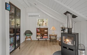 A loft-style room with a wooden floor, a bookshelf, a chair, a table with a lamp, a fireplace, and a window with a view of trees. - 3 bedroom cozy home in Toftlund (Toftlund)