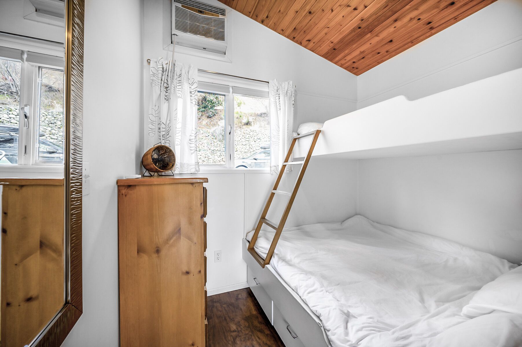 A compact bedroom with a bunk bed, a wooden dresser, a radiator, and a window with curtains.
