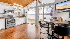 A modern kitchen with stainless steel appliances, a dining area with a round table and black chairs, and a large window offering a view of a lake and mountains.