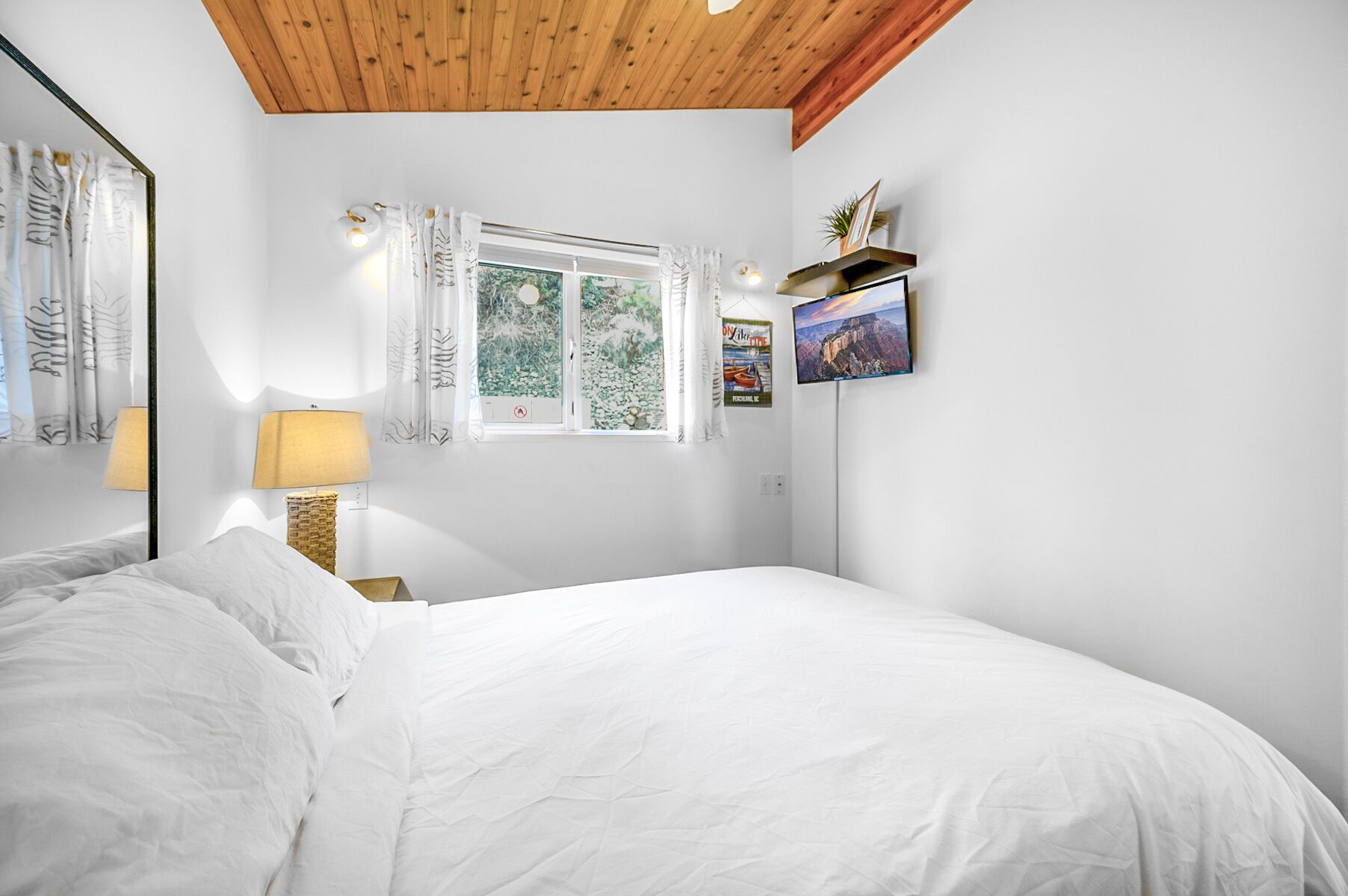 A bedroom with a bed, a window with curtains, a lamp, and a wall-mounted shelf with a plant and a framed picture.