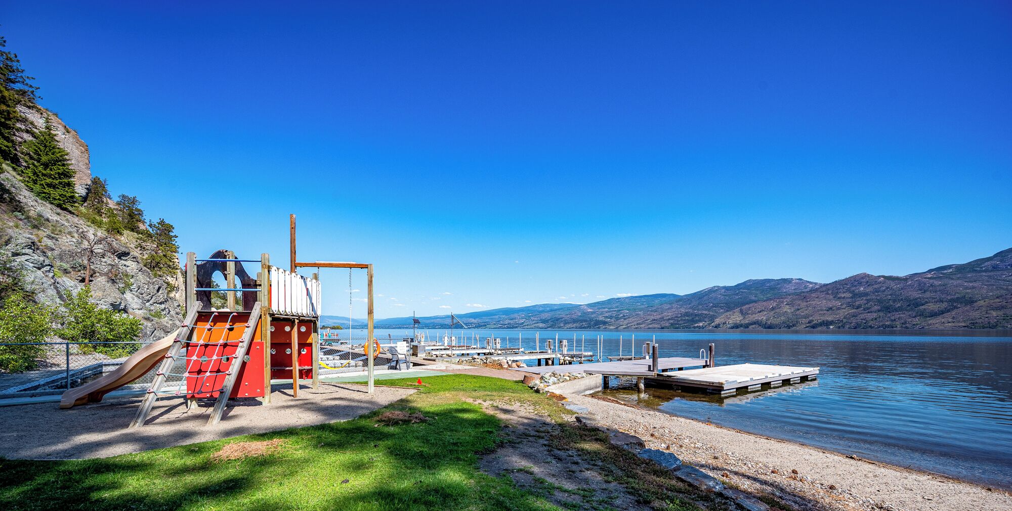 A lakeside playground with a slide and climbing structure, a dock extending into the water, and a mountainous backdrop.