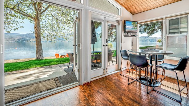 A room with large glass doors leading to a lakeside patio, a dining area with chairs and a table, and a wooden ceiling.