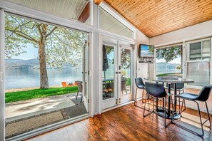 Ein Zimmer mit großen Glasfenstern, die auf eine Terrasse am See führen, einem Essbereich mit Stühlen und Tisch sowie einer Holzbalkendecke.
