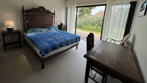 Une chambre à coucher avec un grand lit, deux tables de chevet avec des lampes, une chaise en bois, un lustre et une vue sur un jardin, accessible par une porte-fenêtre coulissante.