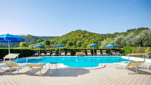 Ein Poolbereich mit Liegestühlen und Sonnenschirmen, umgeben von Grünflächen und Hügeln.