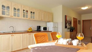 Una cocina con gabinetes blancos, un microondas y un lavamanos. Hay dos sillas de mimbre y una mesa con un juego de vasos de zumo de naranja.