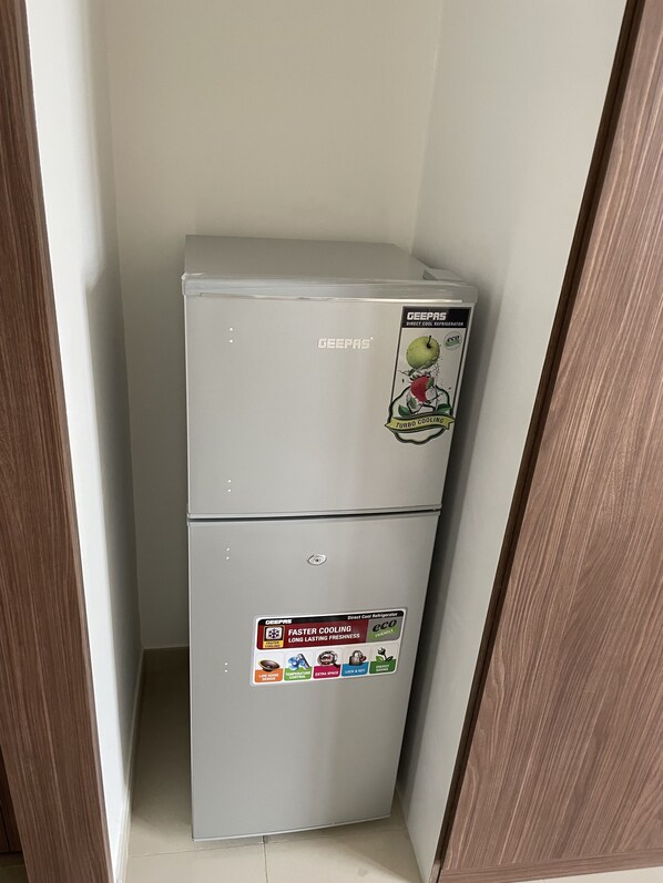 A GEEPAS direct cool refrigerator in a corner of a room with wooden flooring and white walls.