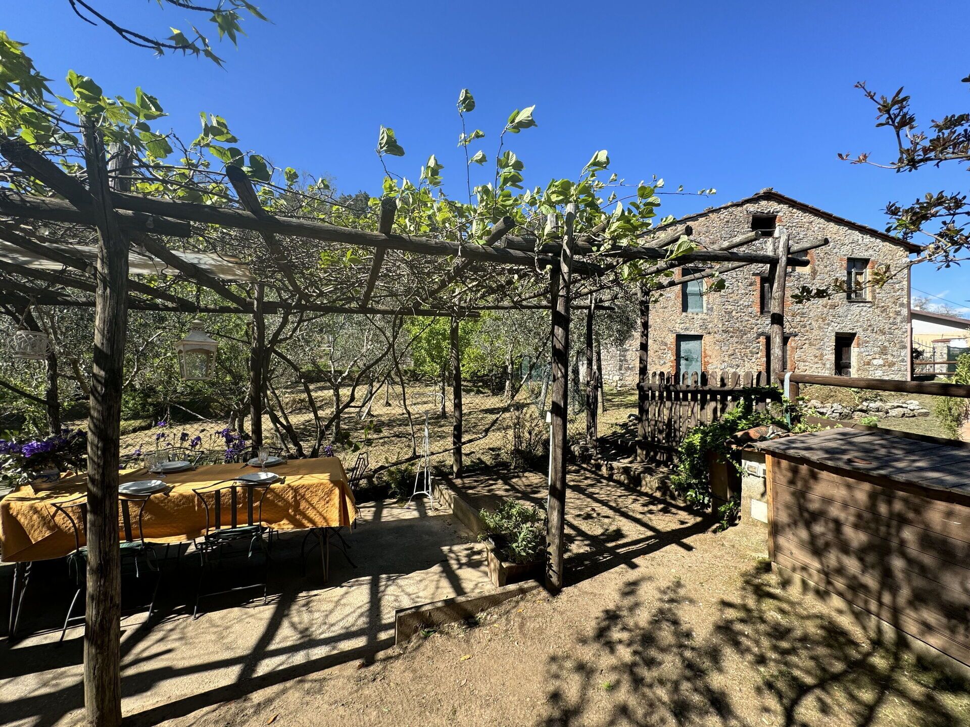 An outdoor dining area with tables covered in yellow tablecloths, a pergola structure, and a stone building in the background.