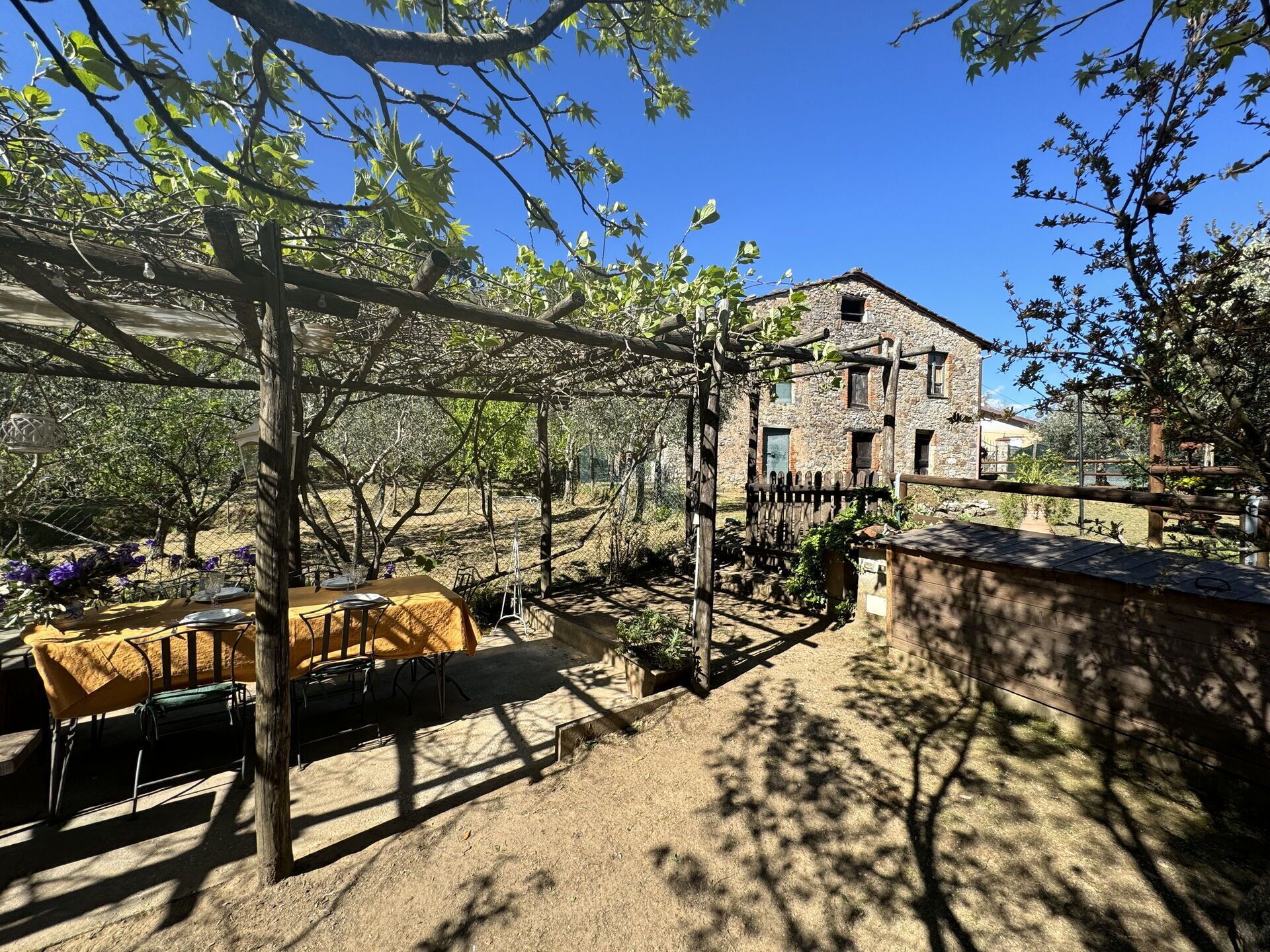 Un área de comedor al aire libre con mesas dispuestas bajo una pérgola, rodeada de árboles y un edificio de piedra al fondo.