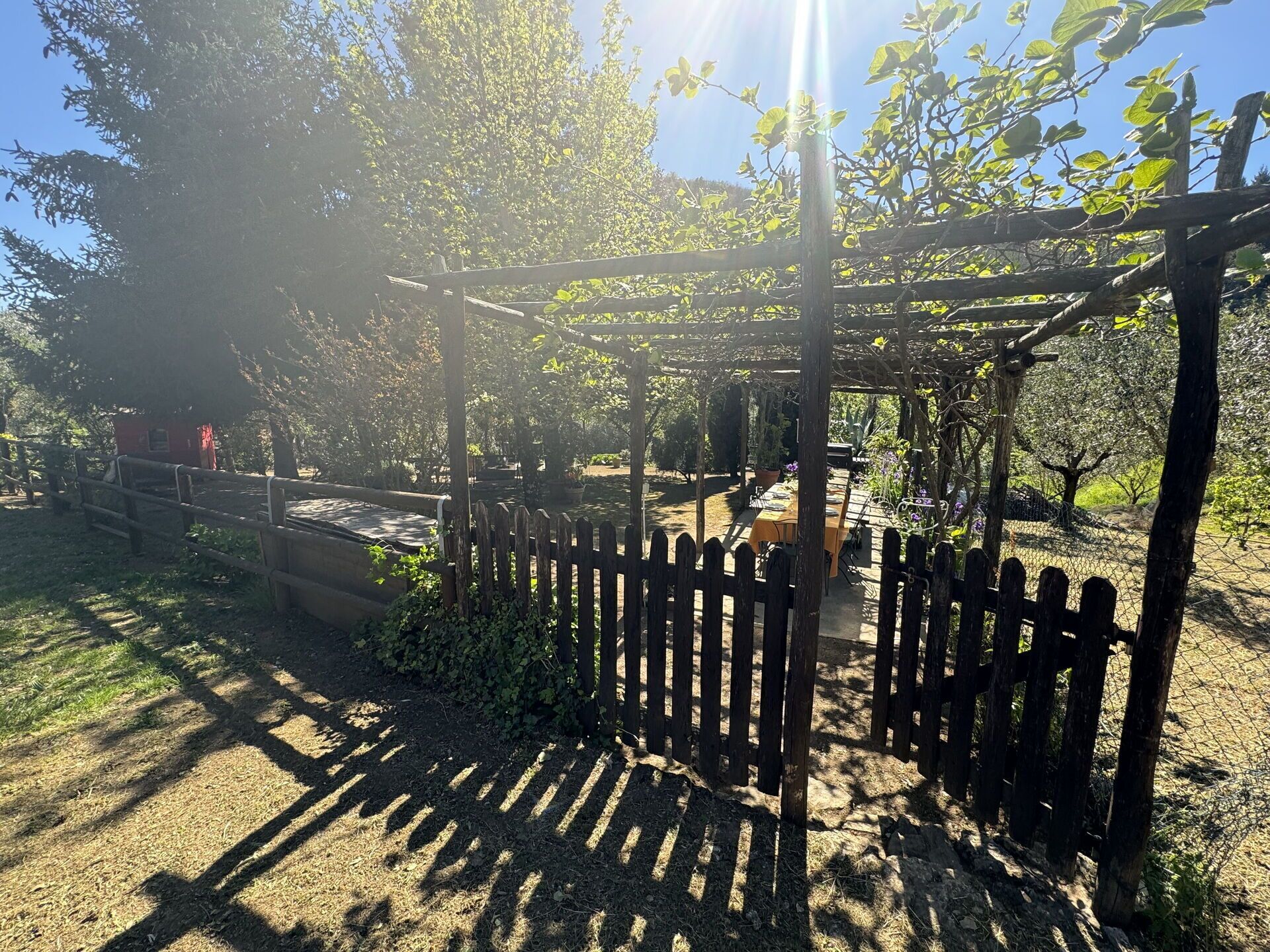 A wooden pergola structure with a table and chairs underneath, surrounded by a wooden fence and greenery.