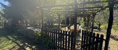 A wooden pergola structure with a table and chairs underneath, surrounded by a wooden fence and greenery.