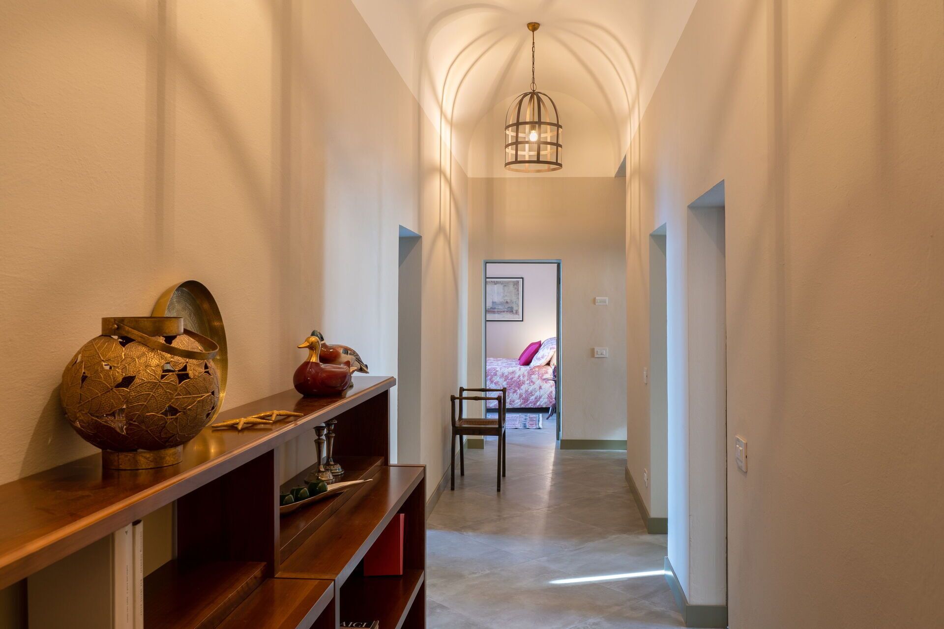 Un pasillo con una consola de madera, un globo decorativo y un cuadro enmarcado a la izquierda. Una silla está apoyada en la pared, y se ve una cama con un cojín rosa al fondo.