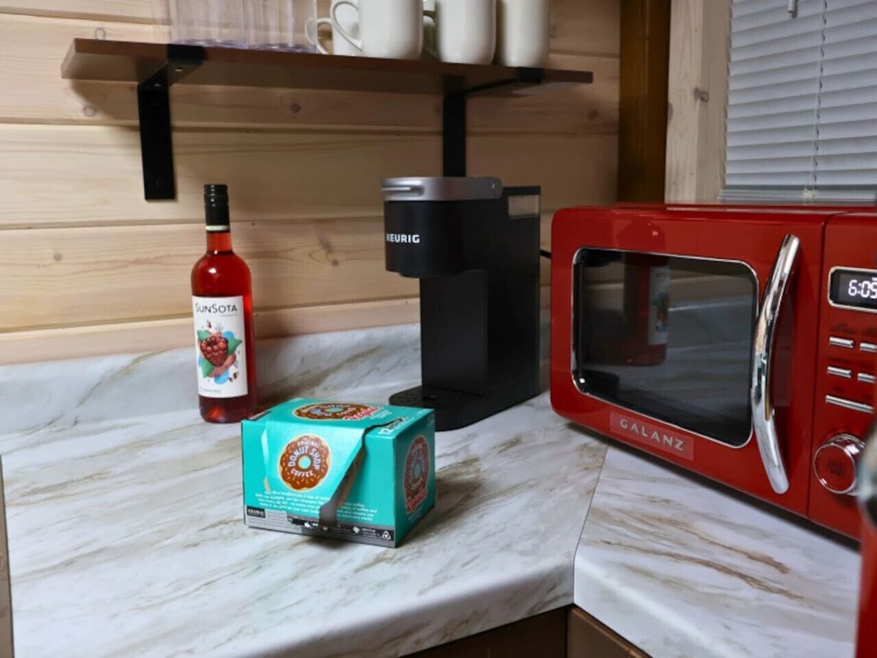 Une cuisine avec un micro-ondes rouge, une machine à café noire, une bouteille de vin et une boîte de café.