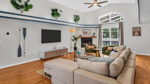 A spacious living room with a large sofa, a fireplace, a ceiling fan, and a TV mounted on the wall.