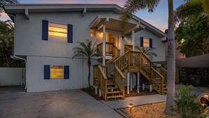 A two-story house with a wooden staircase, a covered porch, and a small garden area with a palm tree.