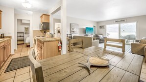 A modern kitchen with wooden cabinets, a dining area with a wooden table and chairs, a living room with a sofa and a TV, and a large window with a view of the outdoors. - Spacious Lakefront Condo w Amazing View (West Kelowna)