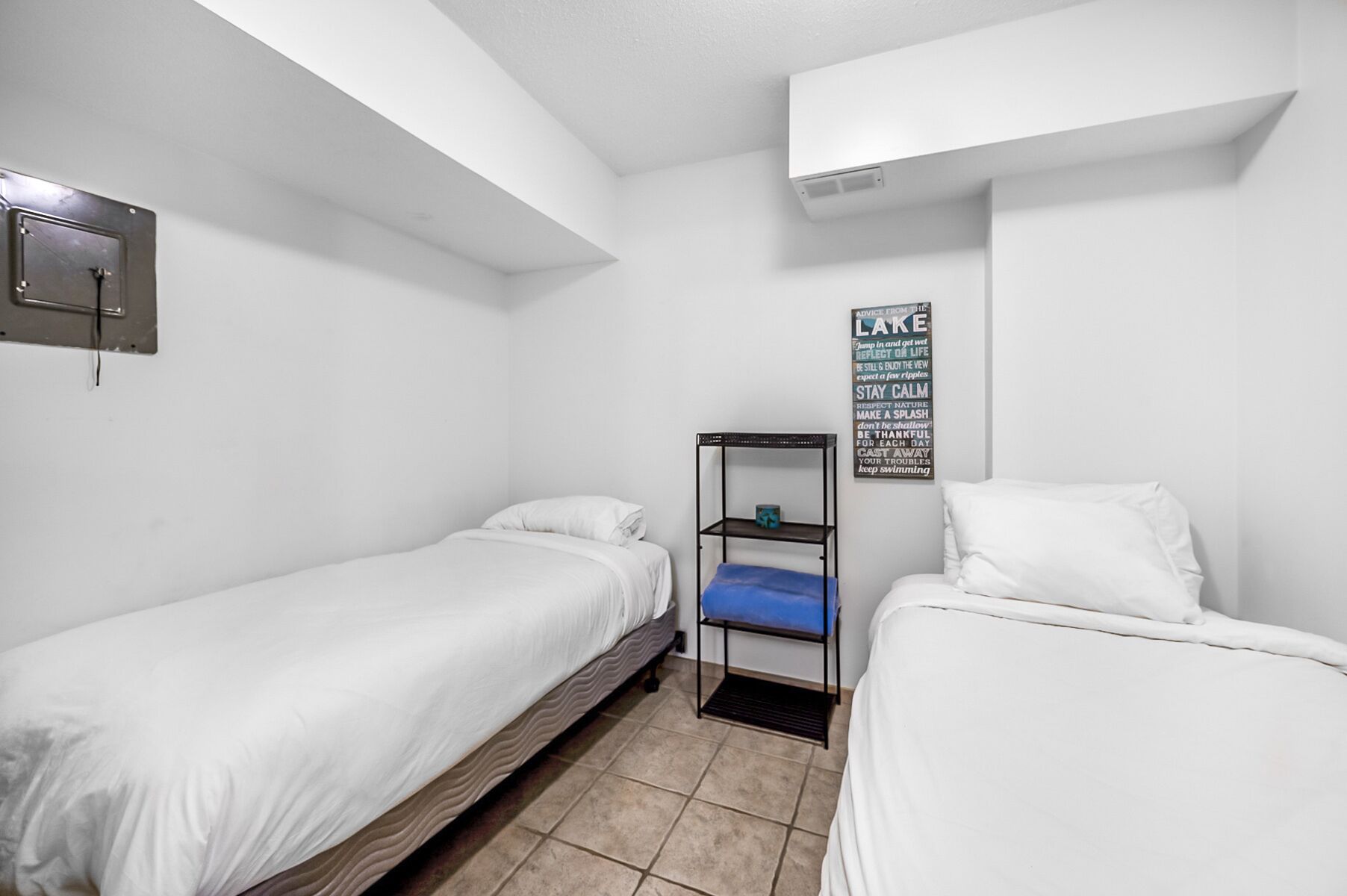 A small, clean room with two single beds, a shelf unit, and a motivational poster on the wall.