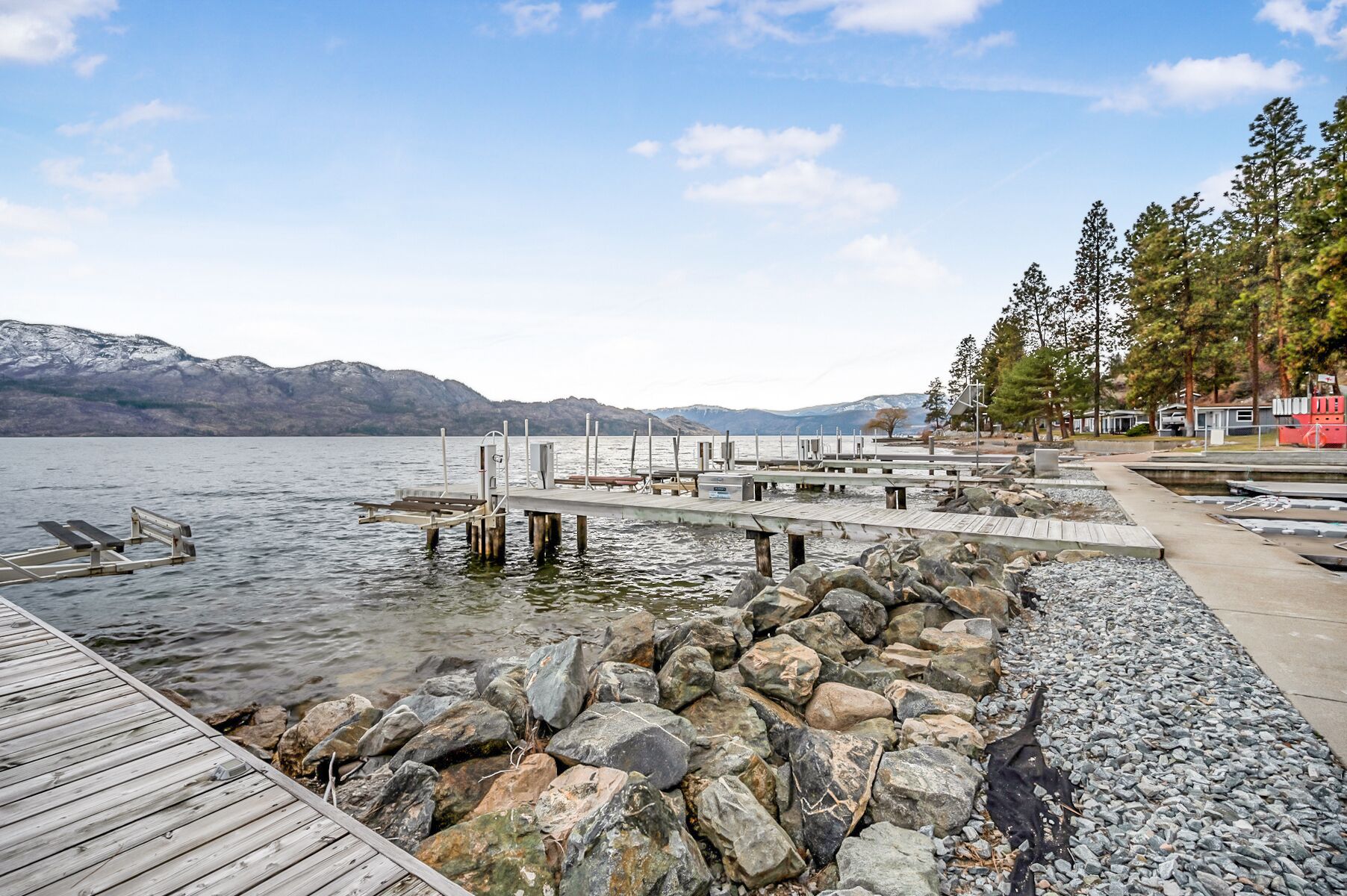 A lakeside dock with a rocky shoreline, a wooden pier extending into the water, and a backdrop of mountains and trees.