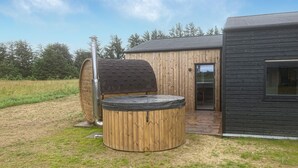 A wooden outdoor hot tub with a hexagonal cover, a wooden deck, and a modern building with a large window.