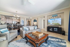 A spacious living room with a large television, a dining area with a chandelier, and a kitchen in the background. - Indigo West Oceanfront Condo | Resort Amenities | My Beach Getaways (Pensacola)