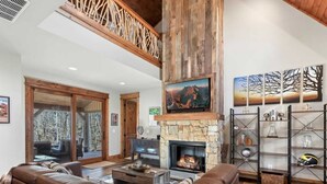 A spacious living room with a stone fireplace, leather sofas, a wooden ceiling, and a large window overlooking a forest.