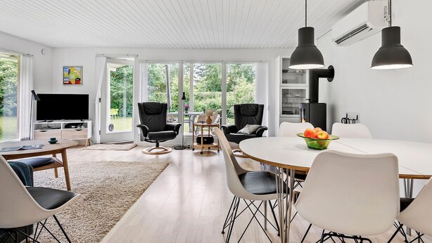 Een moderne woonkamer met een televisie, twee zwarte stoelen, een eettafel met een schaal fruit en een groot raam met uitzicht op groen.