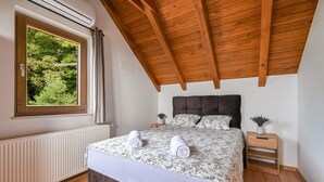 Een slaapkamer met een houten plafond, een groot raam met gordijnen, een radiator, een bed met bloemenbeddengoed, twee nachtkastjes met planten en een witte radiator.