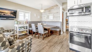 Una cocina moderna con horno empotrado y microondas, un comedor con mesa y sillas de madera, y una zona de estar acogedora con cojines estampados.