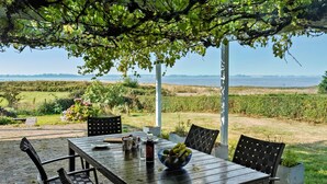 Uma área de refeições coberta ao ar livre, com mesa e cadeiras, uma tigela de frutas e vista para um vinhedo e um corpo de água ao fundo.