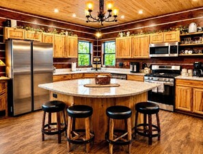 A modern kitchen with wooden cabinets, stainless steel appliances, a central island with stools, and a chandelier.