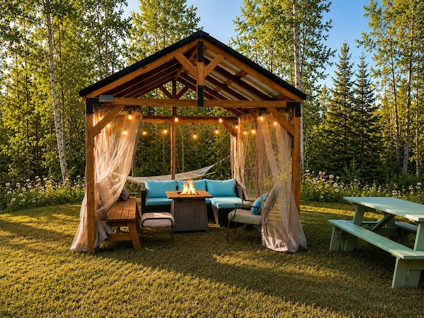 A wooden gazebo with a fire pit, cushioned seating, and a picnic table in a forested area.